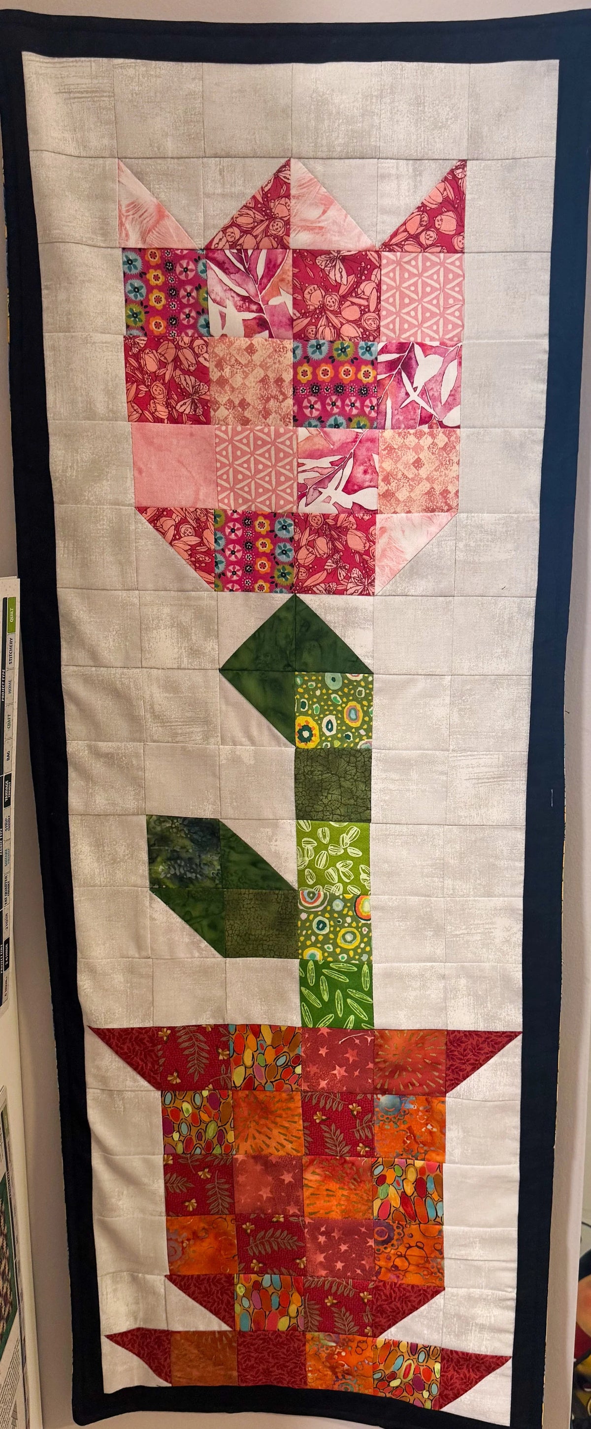 Beige fabric patchwork wall hanging with red flower, green stems, and black border