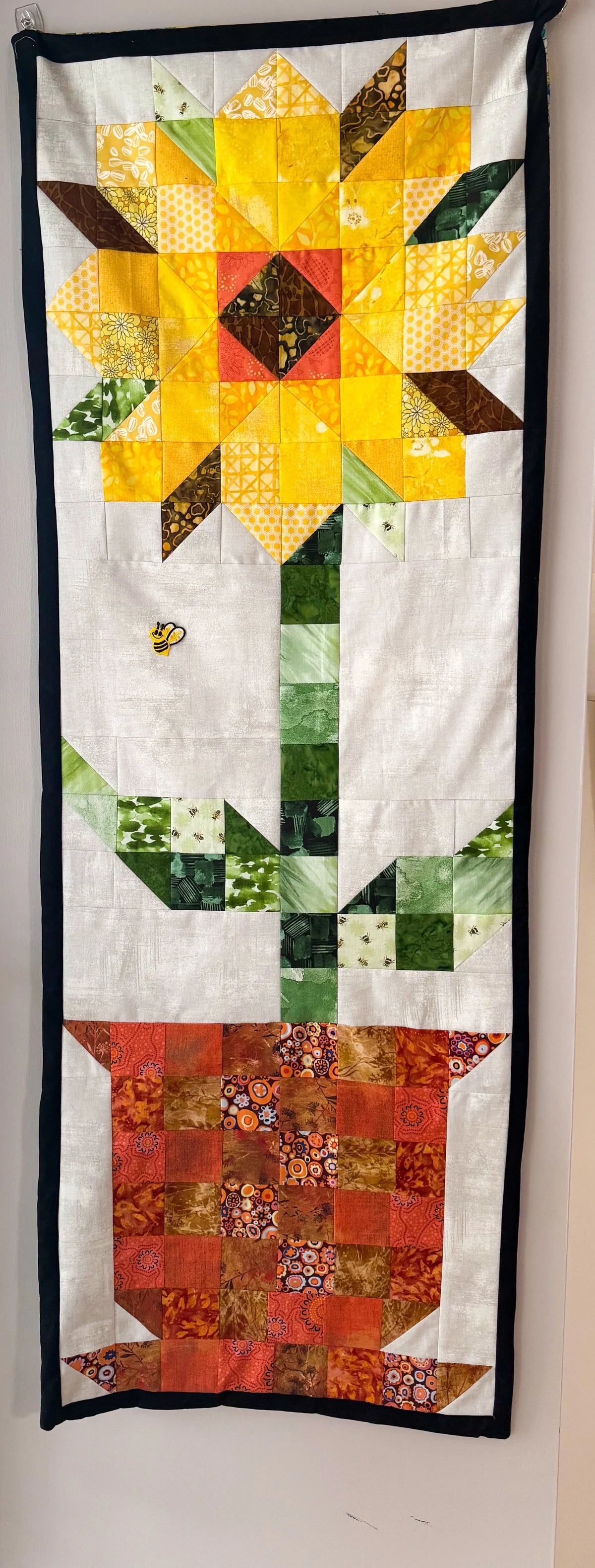 White fabric patchwork wall hanging with yellow flower top, green leaves, red base, black border