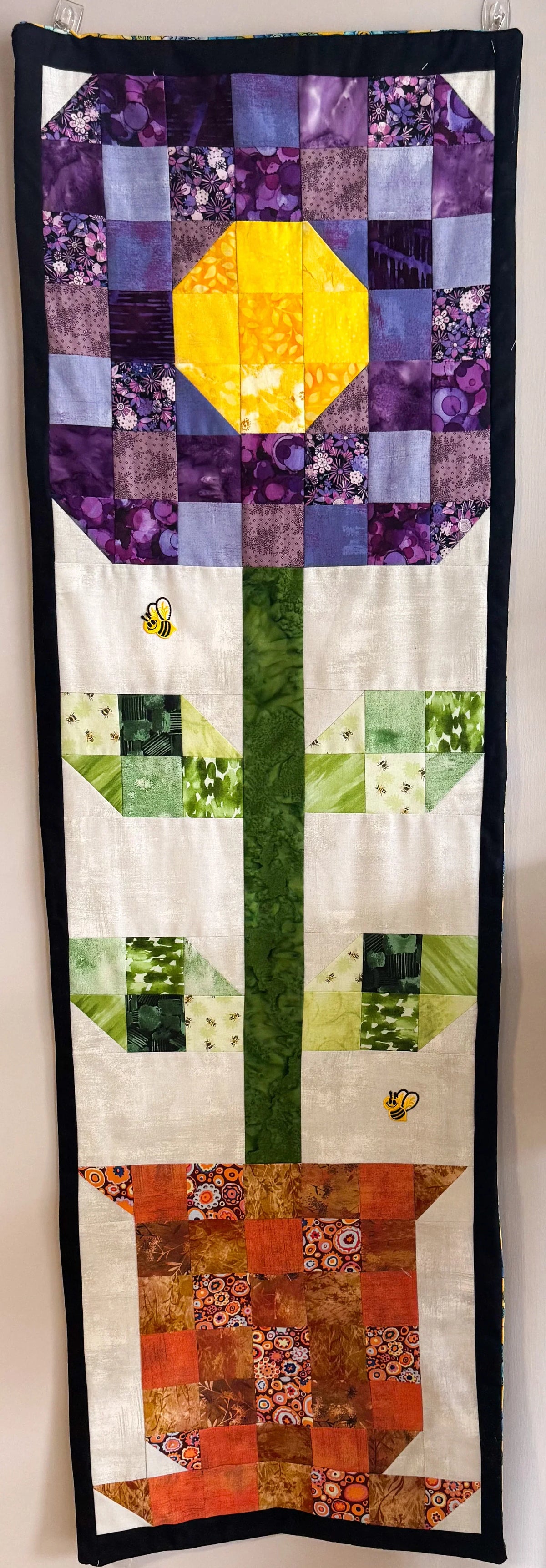 Beige and purple patchwork wall hanging with yellow center flower, green accents, black border