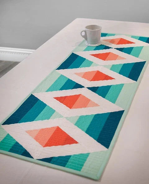 Quilted table runner with teal and green textured blocks and coral diamond accents on white