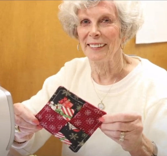 An elderly woman holding up a small, handmade Christmas-themed quilted coaster.